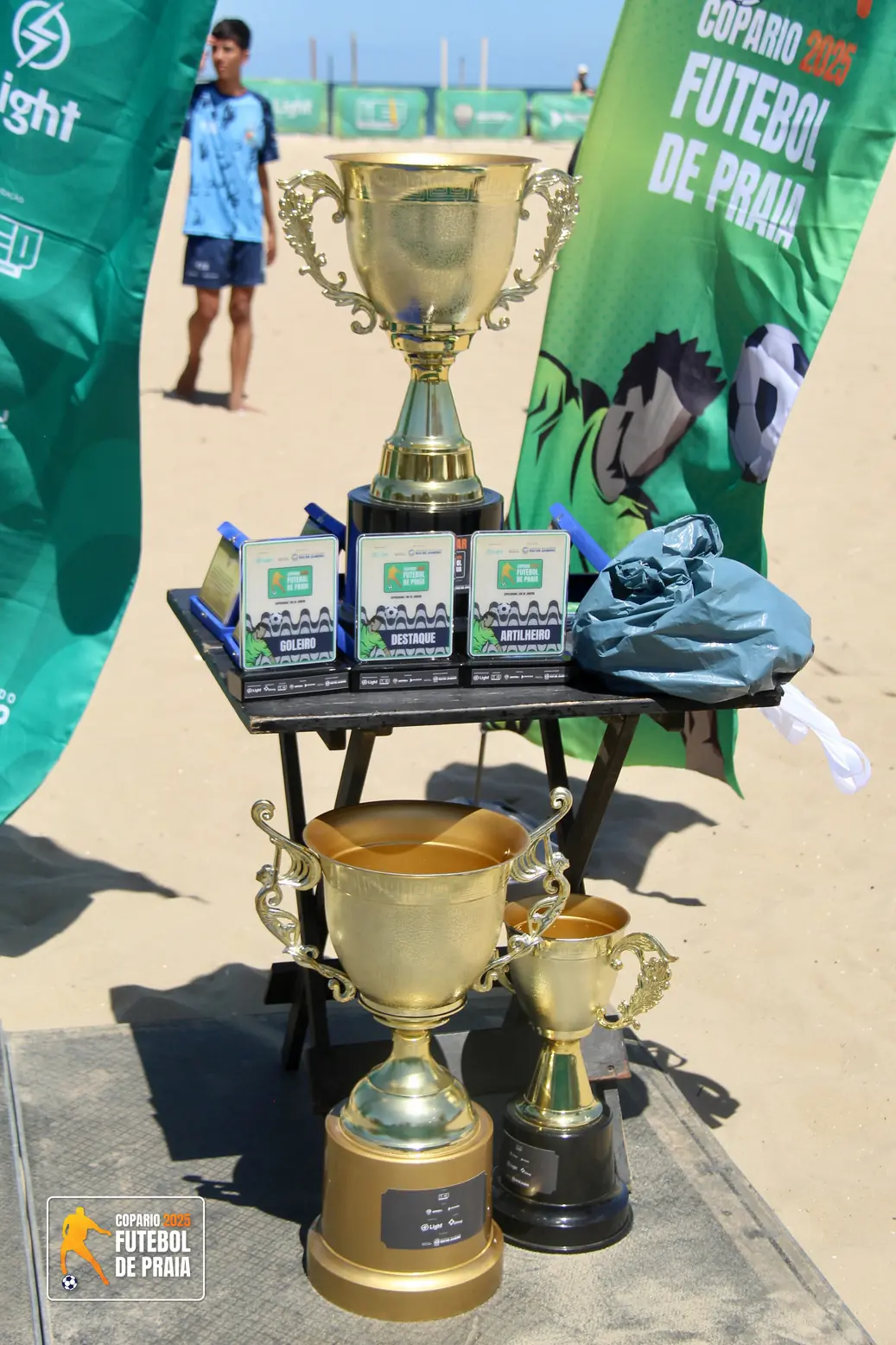 Mesa com troféus dourados e medalhas de premiação sobre a areia