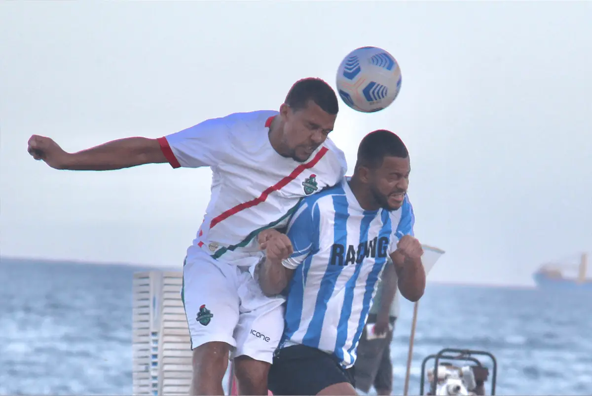 Dois jogadores sobem para cabecear a bola, com o mar ao fundo