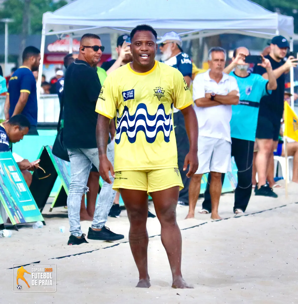 Jogador de camisa amarela sorri diante das tendas do torneio