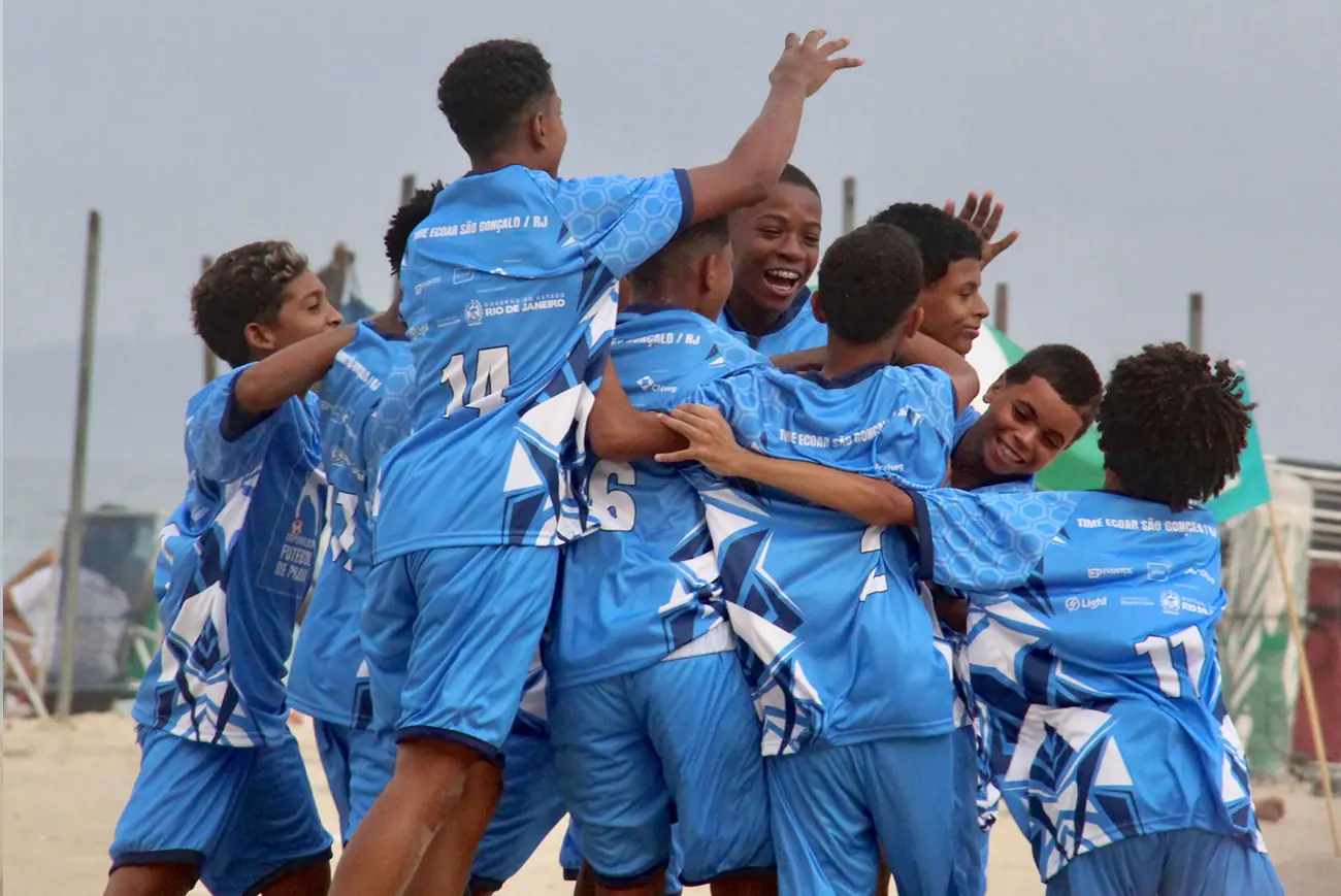 Meninos de camisa azul se abraçam e comemoram um gol na areia